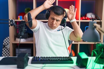 Young hispanic man playing video games doing bunny ears gesture with hands palms looking cynical...