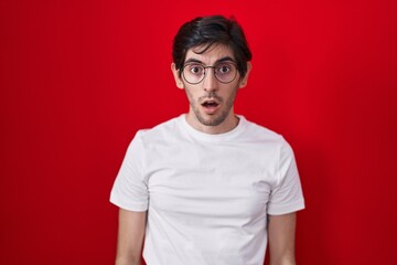 Young hispanic man standing over red background afraid and shocked with surprise and amazed expression, fear and excited face.