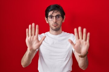 Young hispanic man standing over red background moving away hands palms showing refusal and denial with afraid and disgusting expression. stop and forbidden.