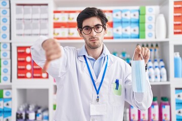 Young hispanic man working at pharmacy drugstore holding safety mask with angry face, negative sign showing dislike with thumbs down, rejection concept