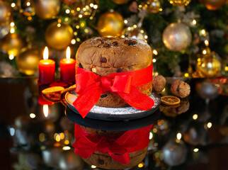 Italian Christmas cake Panettone with a red bow on a black glass against the background of a decorated Christmas tree