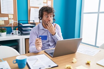 Hispanic young man working at the office doing online shopping covering mouth with hand, shocked...