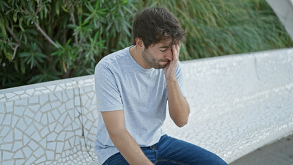 Serious young hispanic man, rocking a blond beard, combating stress sitting on city park bench. his...