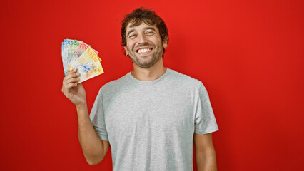 Happy young man with blond beard proudly holding up swiss franc banknotes, financial triumph writ large on his face, depicted in an isolated, vivacious red background portrait! - obrazy, fototapety, plakaty
