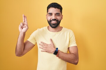 Hispanic man with beard standing over yellow background smiling swearing with hand on chest and...