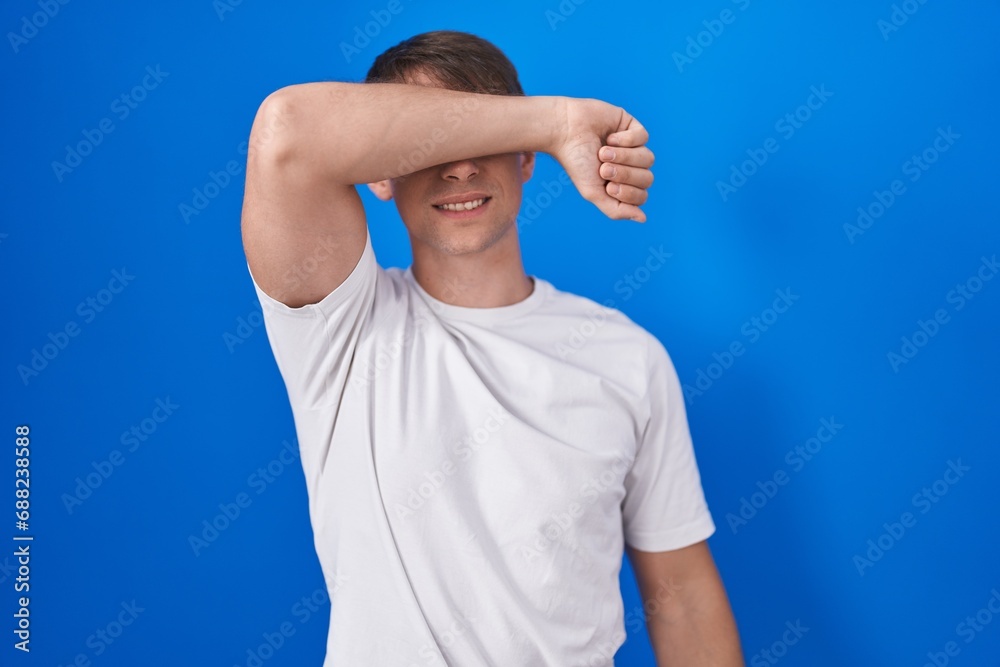 Sticker Caucasian blond man standing over blue background covering eyes with arm smiling cheerful and funny. blind concept.