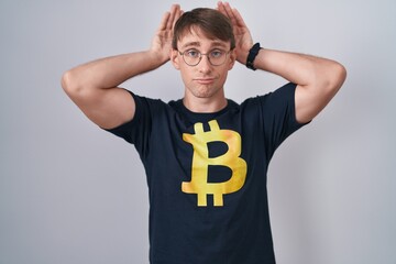 Caucasian blond man wearing bitcoin t shirt doing bunny ears gesture with hands palms looking...
