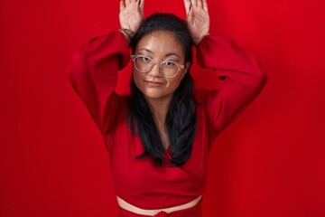Asian young woman standing over red background doing bunny ears gesture with hands palms looking cynical and skeptical. easter rabbit concept.