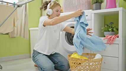 Young blonde woman listening to music holding clothes of basket at laundry room