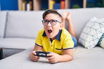 Young hispanic kid playing video game holding controller on the sofa angry and mad screaming...