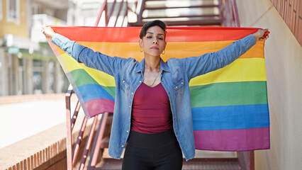 Young beautiful hispanic woman standing with serious expression holding rainbow flag at street