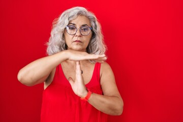 Middle age woman with grey hair standing over red background doing time out gesture with hands,...