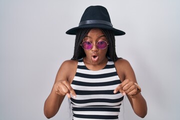 Young african american with braids wearing hat and sunglasses pointing down with fingers showing advertisement, surprised face and open mouth