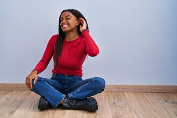 Young african american with braids sitting on the floor at home smiling with hand over ear...