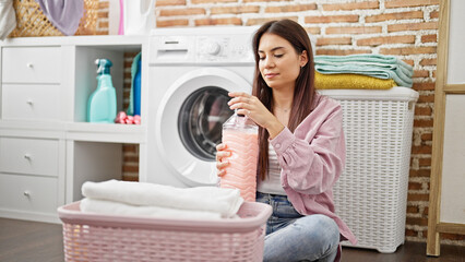 Young beautiful hispanic woman washing clothes holding detergent bottle at laundry room