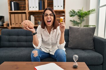 Young hispanic woman working on depression holding pills angry and mad screaming frustrated and furious, shouting with anger looking up.