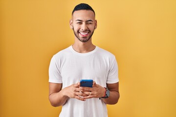 Young hispanic man using smartphone typing message winking looking at the camera with sexy...