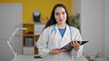 Young beautiful hispanic woman doctor holding clipboard smiling at the clinic