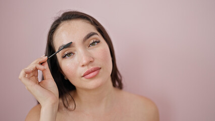Young beautiful hispanic woman applying make up on eyelashes over isolated pink background