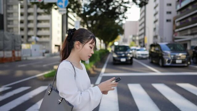 愛知県名古屋市大須商店街周辺の道路沿いでスマートフォンを操作しながら歩く若い日本人女性 Young Japanese woman walking while operating a smartphone along a road near the Osu shopping district in Nagoya City, Aichi Prefecture, Japan.
