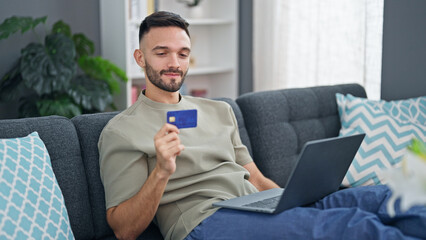 Young hispanic man shopping with laptop and credit card sitting on sofa at home
