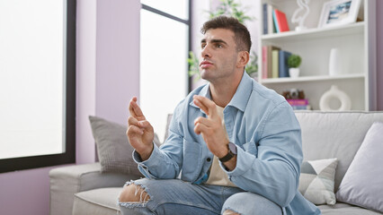 Hopeful young hispanic man sitting at home, crossing fingers for luck while watching a movie on his...