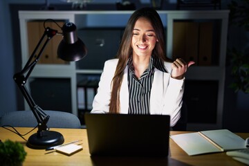 Young brunette woman working at the office at night with laptop pointing to the back behind with...