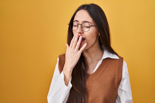 Young brunette woman standing over yellow background wearing glasses bored yawning tired covering mouth with hand. restless and sleepiness.
