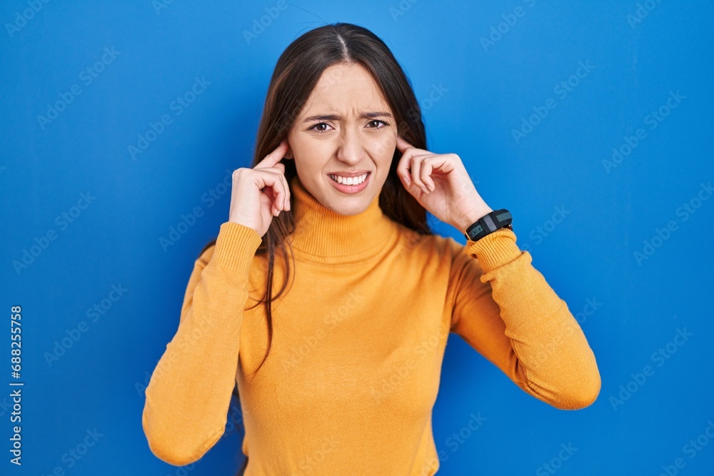 Wall mural Young brunette woman standing over blue background covering ears with fingers with annoyed expression for the noise of loud music. deaf concept.