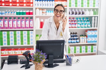 Young beautiful hispanic woman pharmacist talking on telephone using computer at pharmacy