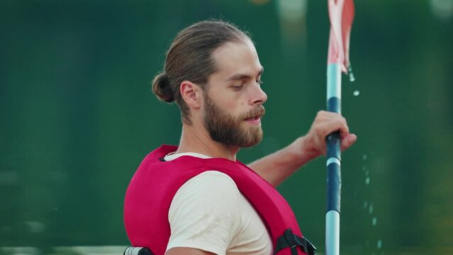 Side view of handsome Caucasian male with beard and man bun hairstyle. Wearing red safety jacket and rowing in river with focus. Carefully trying to navigate in water. Learning something new.