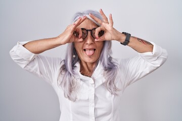 Middle age woman with tattoos wearing glasses standing over white background doing ok gesture like binoculars sticking tongue out, eyes looking through fingers. crazy expression.