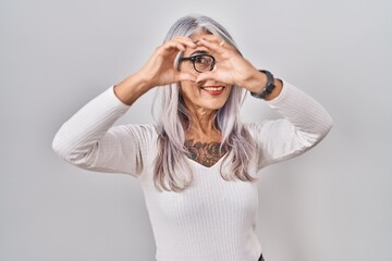 Middle age woman with grey hair standing over white background doing heart shape with hand and fingers smiling looking through sign