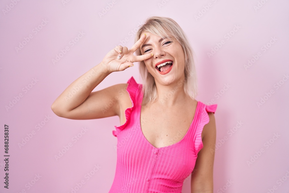 Poster young caucasian woman standing over pink background doing peace symbol with fingers over face, smili