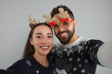 Happy young couple in Christmas sweaters, reindeer headband and funny glasses taking selfie on grey...