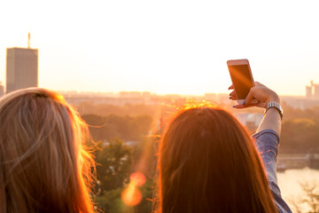 Two girls taking selfie during sunset near the river. Shot from behind.