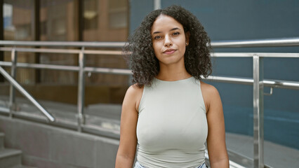 Attractive young hispanic woman, with curly hair, displaying a serious expression while standing on a sunlit city street, living a relaxed yet cool, urban lifestyle.
