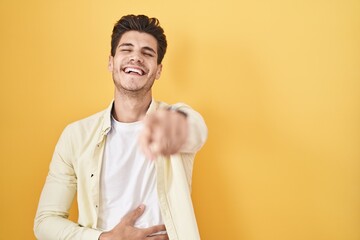 Young hispanic man standing over yellow background laughing at you, pointing finger to the camera...