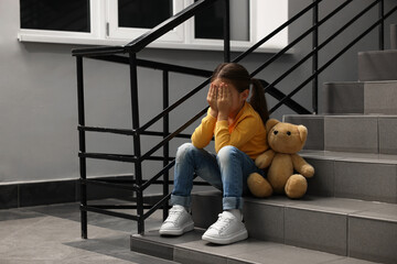 Child abuse. Upset little girl with teddy bear sitting on stairs indoors