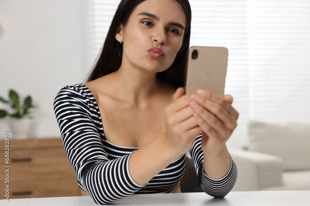 Sticker Young woman having video chat via smartphone and sending air kiss at table in room