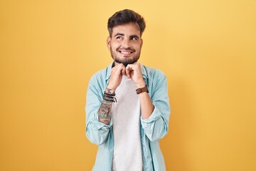 Young hispanic man with tattoos standing over yellow background laughing nervous and excited with...