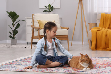 Little girl petting cute ginger cat on carpet at home