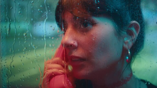 Beautiful girl making a call and talking on payphone inside wet phone booth at rainy day in the city, street traffic in the background