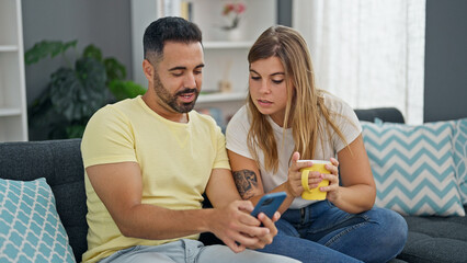 Man and woman couple drinking coffee using smartphone sitting on sofa at home
