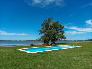 Lawn terrace with swimming pool and deckchairs under a tree for relaxation at the shore of a lake