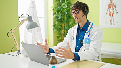 Young hispanic man doctor doing video call with laptop and headphones at the clinic