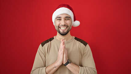 Young arab man wearing christmas hat standing with hands together over isolated red background