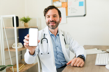 Cheerful caucasian doctor smiling using a medical app on his smartphone and showing the blank phone...