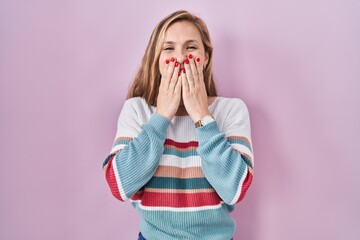 Young blonde woman standing over pink background laughing and embarrassed giggle covering mouth...