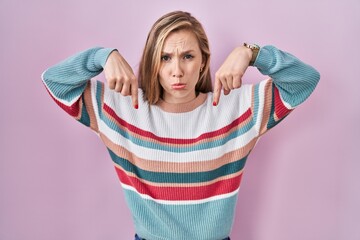 Young blonde woman standing over pink background pointing down looking sad and upset, indicating...